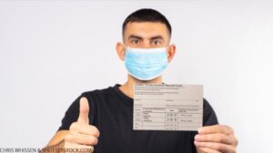 A man in a medical mask holds a coronavirus vaccination card and gestures thumbs up.